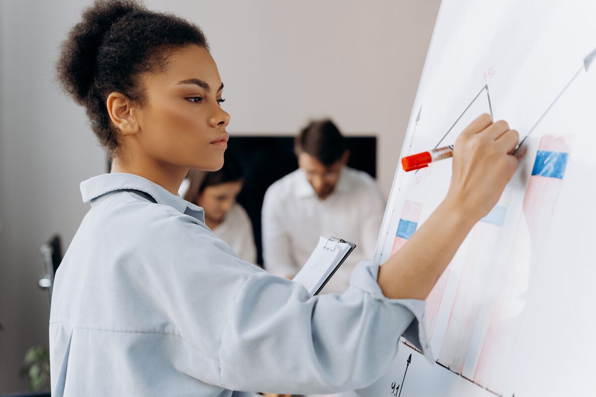 A professional draws on a whiteboard while colleagues work in the background.